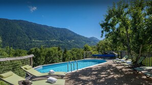Une terrasse en bois avec des chaises longues et une piscine, entourée d’une végétation luxuriante et de montagnes.