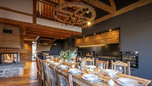 Un coin repas rustique avec une longue table en bois dressée pour le repas, une cheminée en pierre et, au fond, une cuisine moderne.