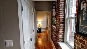 Un couloir avec un plancher en bois, des portes blanches et un mur en briques percé d’une fenêtre.