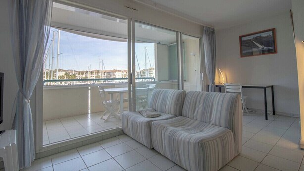 A modern living room with a sofa, a dining table, and a view of a marina through large glass doors. - Apartment With Balcony And View On The Port. (Fréjus)