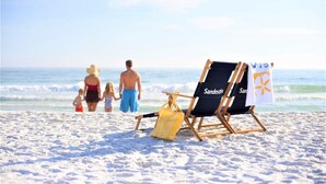 Una playa con una familia caminando hacia el océano, una silla de playa con el nombre "Sandy" y una toalla con un logotipo.