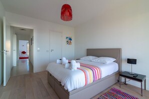 Quarto com cama, mesa de cabeceira com luminária, luminária pendente vermelha, tapete colorido e porta que dá para outro cômodo.