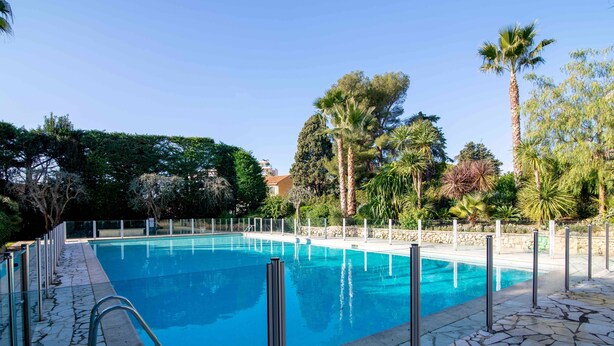 A rectangular swimming pool with a stone-tiled deck, surrounded by a metal fence and various trees. - Apartment In A Residence With Swimming Pool Wifi (Cannes)