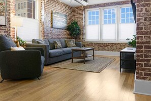 A living room with a brick wall, a grey sofa, a coffee table, and a wooden floor. - Gorgeous 2BR downtown condo near EVERYTHING! (Memphis)