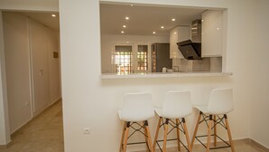 A kitchen with a built-in stove, a sink, and a window with a view of greenery. There are three white bar stools with wooden legs.