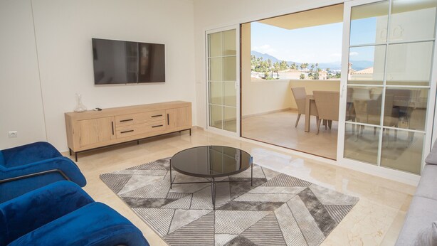 A modern living room with a flat-screen TV, a wooden cabinet, a blue sofa, a patterned rug, and a glass door leading to a balcony with outdoor furniture.