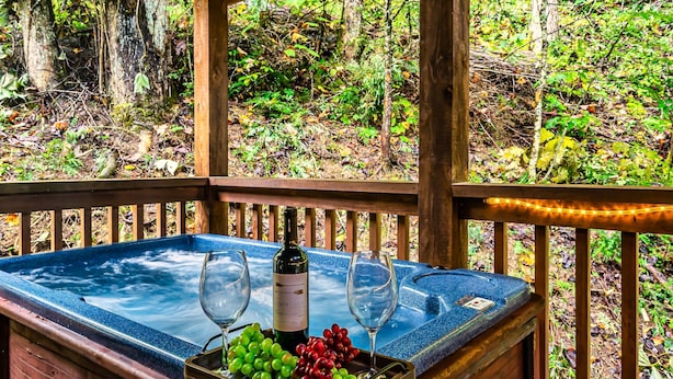 Uma banheira de hidromassagem com uma garrafa de vinho e uvas, rodeada por um deck de madeira e vista para a floresta.