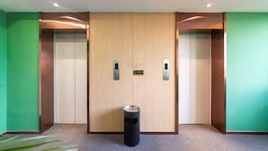 A hotel lobby with a central elevator flanked by green walls and a trash bin in the middle. - Lianjia Holiday Hotel (Jigang Senior High School) (Jinan)