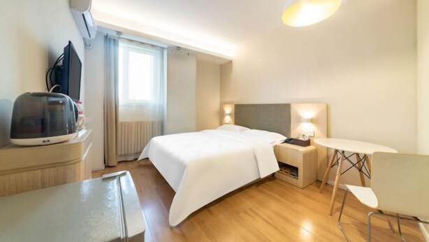 A hotel room with a bed, bedside table, lamp, and a small table with a chair. - Lianjia Holiday Hotel (Jigang Senior High School) (Jinan)