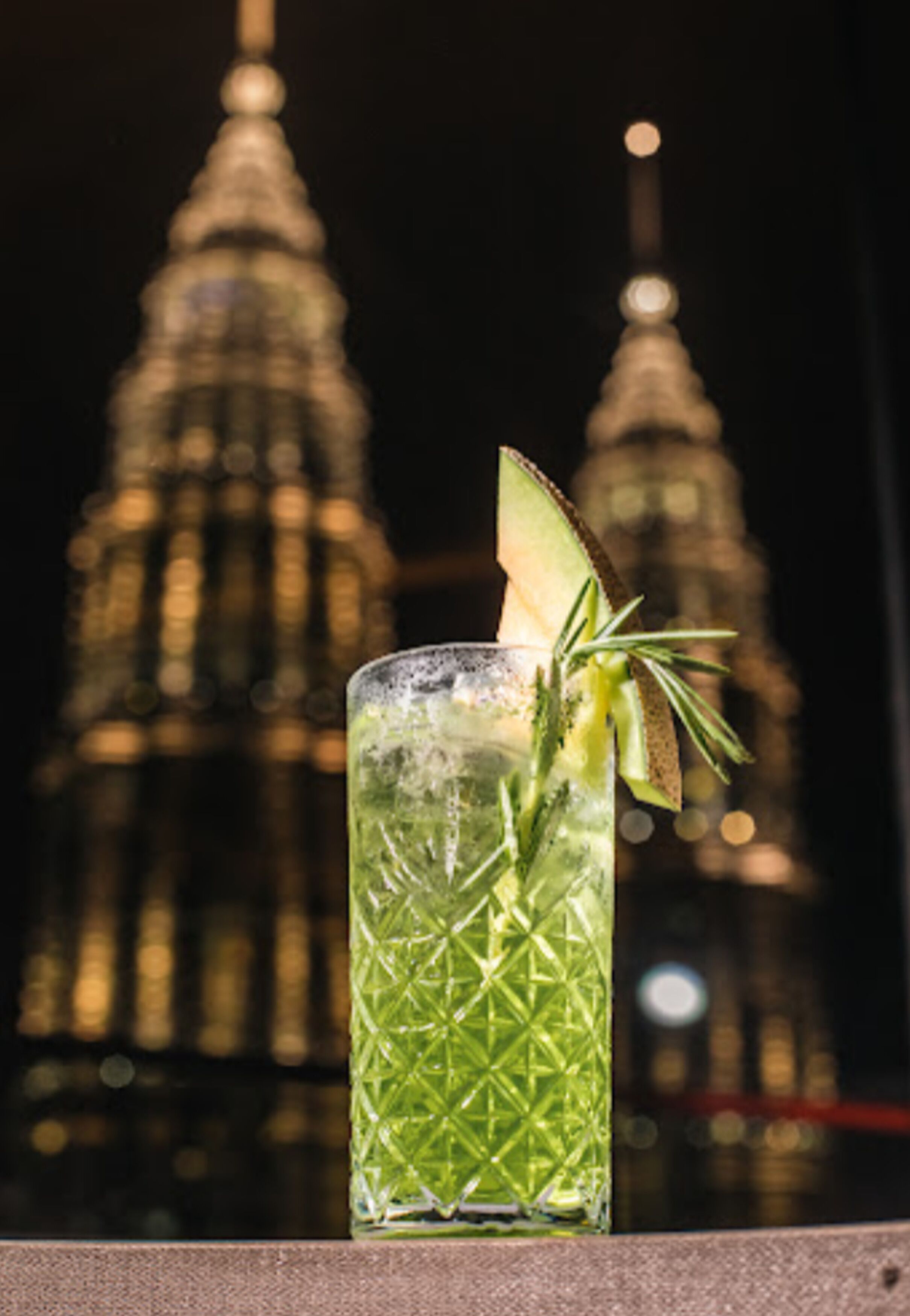 A tall, clear glass with a green drink, garnished with a slice of lime and a sprig of rosemary, set against a blurred background of a building with a pointed top.