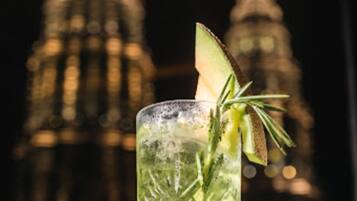 A tall, clear glass with a green drink, garnished with a slice of lime and a sprig of rosemary, set against a blurred background of a building with a pointed top.