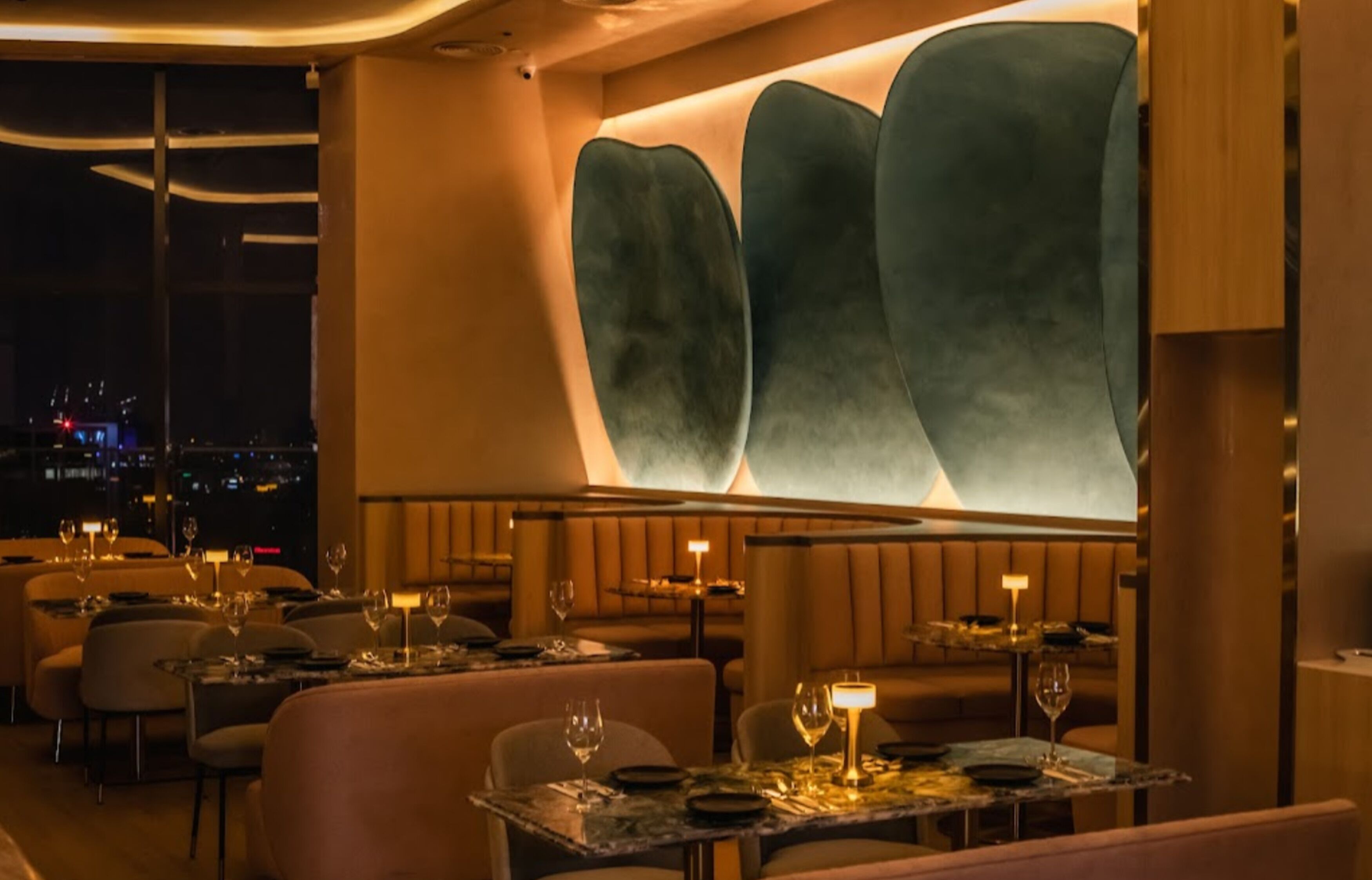 A restaurant with tables set for dining, a large illuminated wall, and a city view.