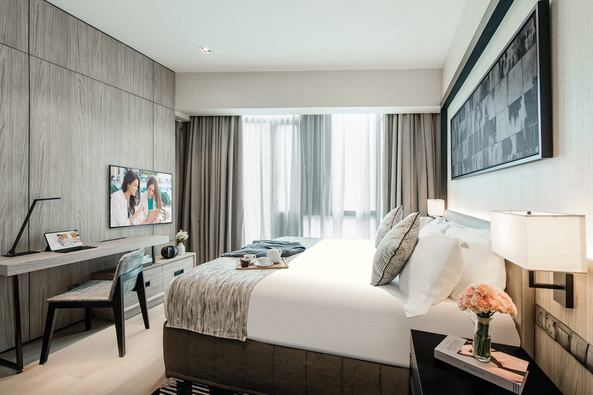 Moderni hotellihuone, jossa on suuri sänky, työpöytä kannettavalla tietokoneella, tuoli, yöpöytä maljakko täynnä kukkia ja suuri ikkuna, jossa on verhot.