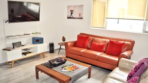 Un salon avec un canapé en cuir, un meuble de rangement blanc pour appareils électroniques, un téléviseur et une fenêtre avec des stores.