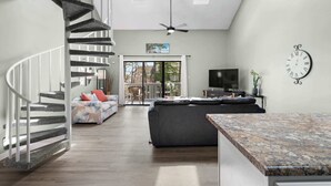 A modern living space with a spiral staircase, a large dark sofa, a flat-screen TV, and a clock on the wall. - Rudy's Reel 'Em Inn By Beach Star (Myrtle Beach)