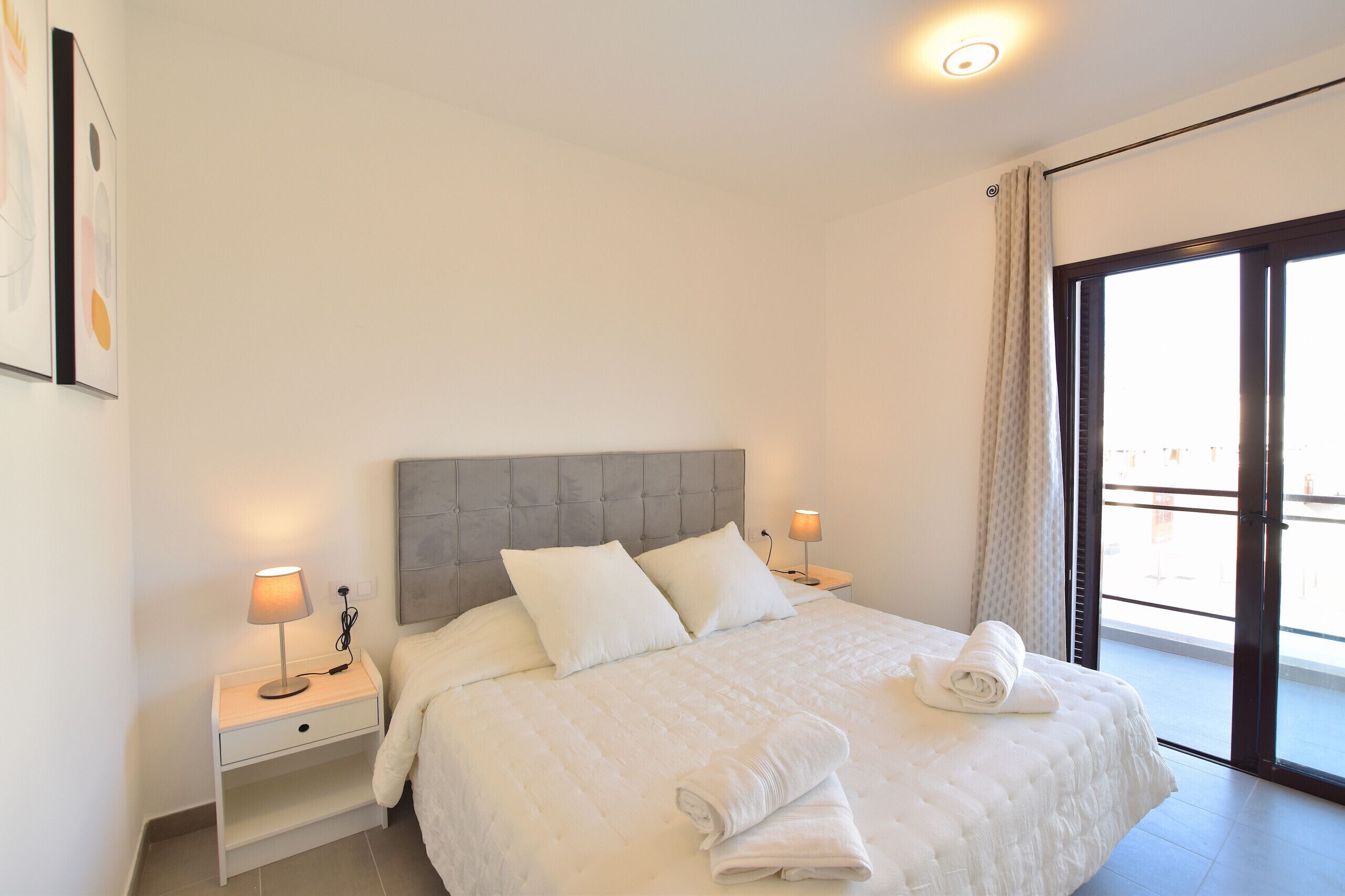 A neatly made bed with white linens, two bedside tables with lamps, a grey headboard, and a view through a glass door.