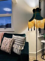 A dark green sofa with patterned cushions, a floor lamp with tassels, and a view of the cityscape through a window.