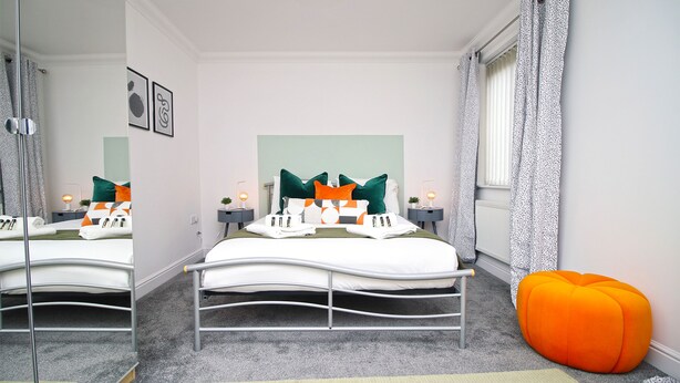 A bedroom with a bed, bedside tables, a mirror, a window with curtains, and a large orange bean bag.