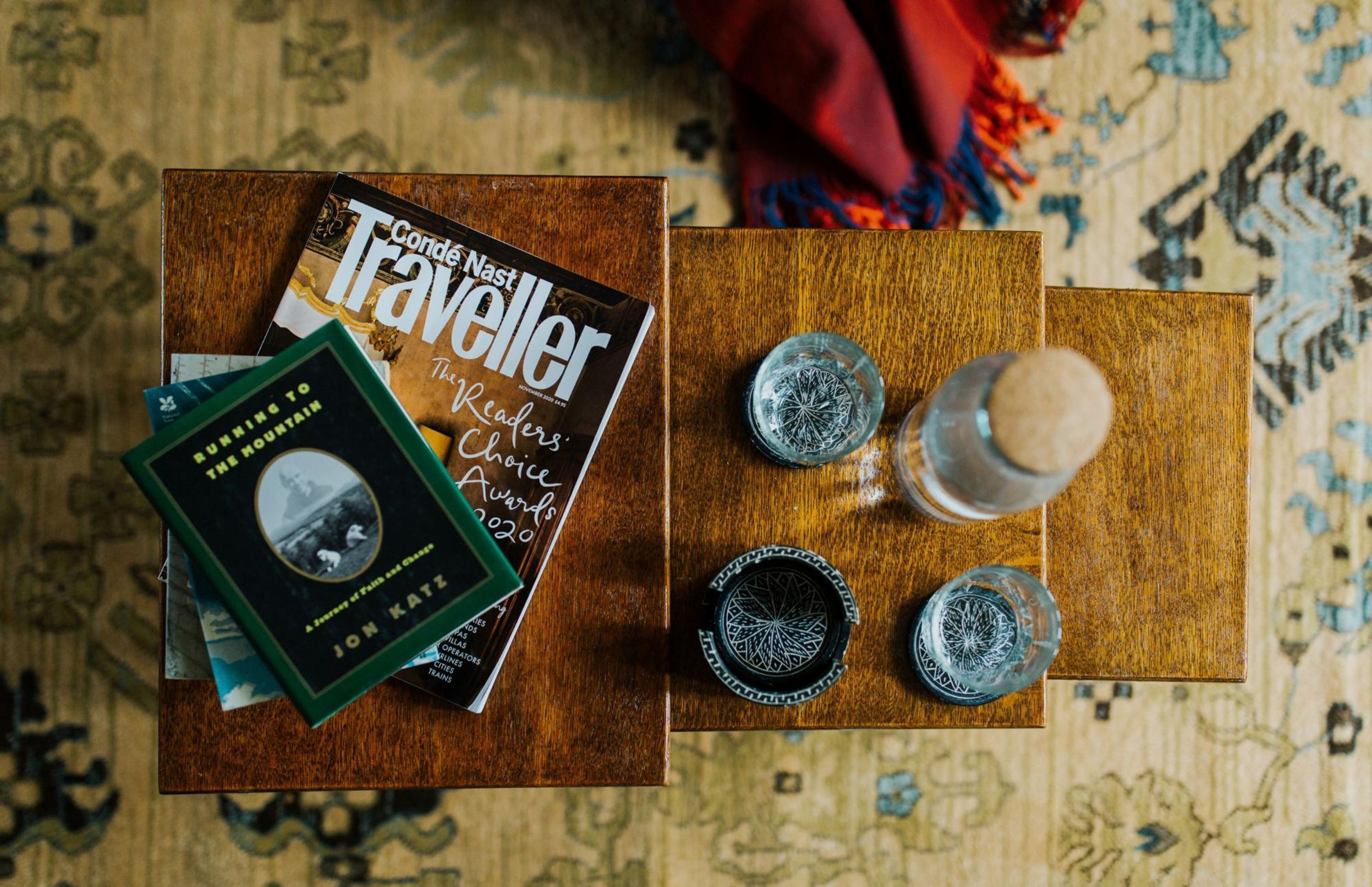 Una mesa de madera con un libro titulado 'Condé Nast Traveller' y otro titulado 'Running to the Mountain'. Hay tres vasos sobre la mesa, uno con un tapón de corcho y dos con diseños intrincados.