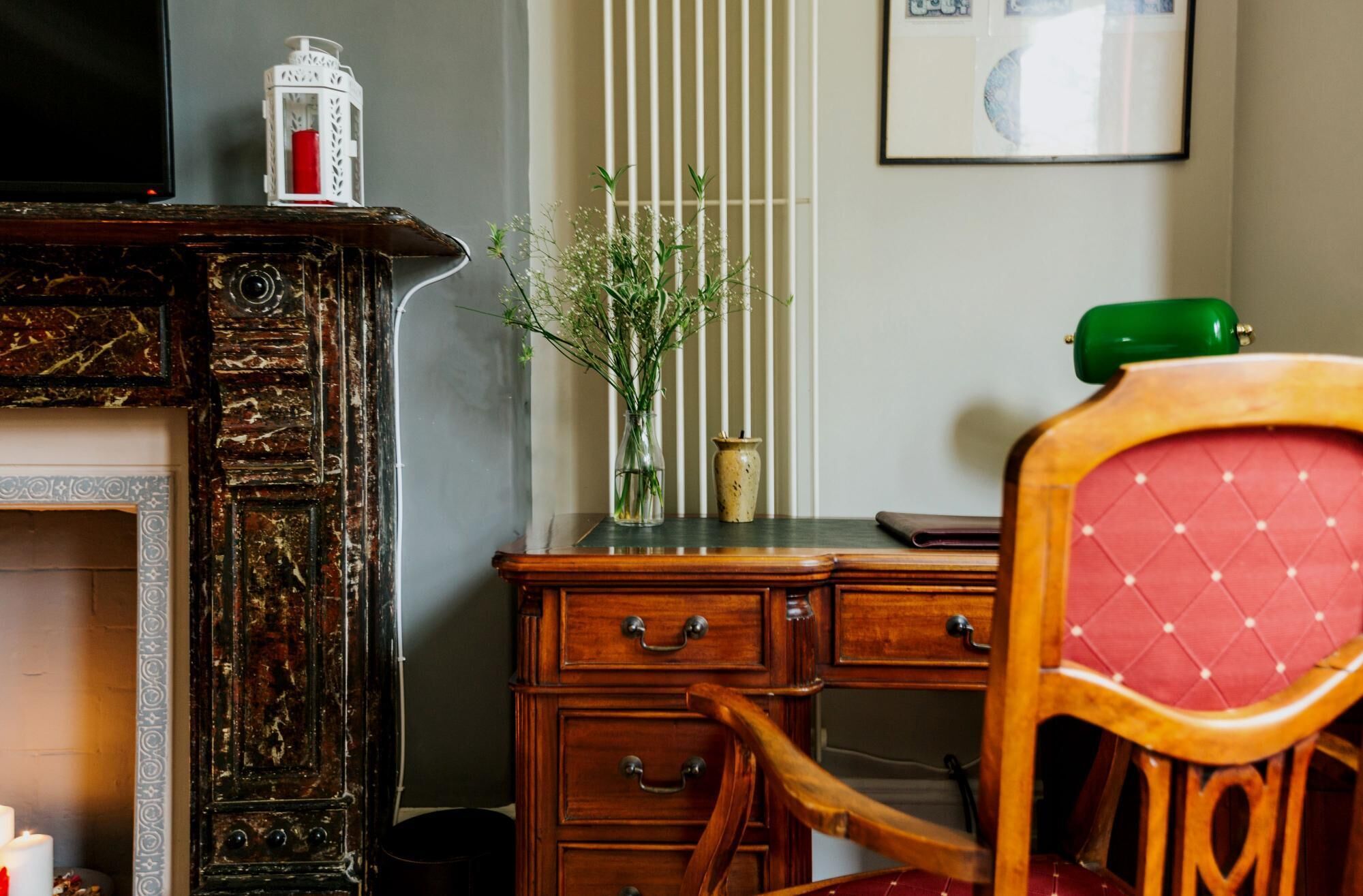 Un escritorio de madera con cajones, un florero con flores, una lámpara verde y una silla con un cojín rojo.