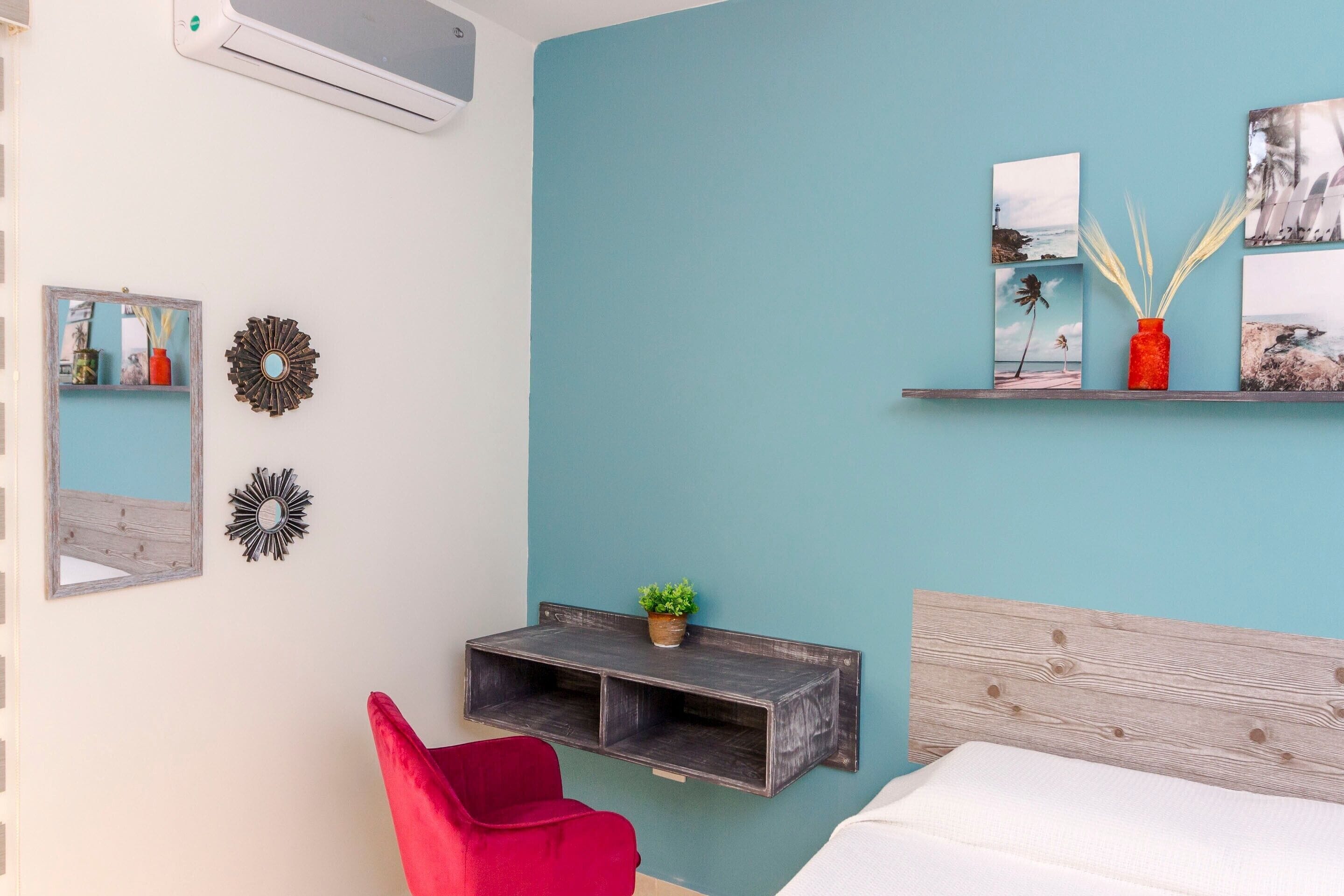 A bedroom with a teal wall, a wooden headboard, a red chair, a shelf with pictures and a vase, and a small plant.