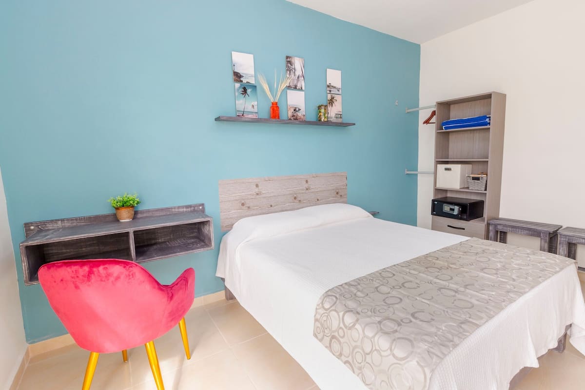 Um quarto com cama, escrivaninha com cadeira, estante com obras de arte e guarda-roupa.