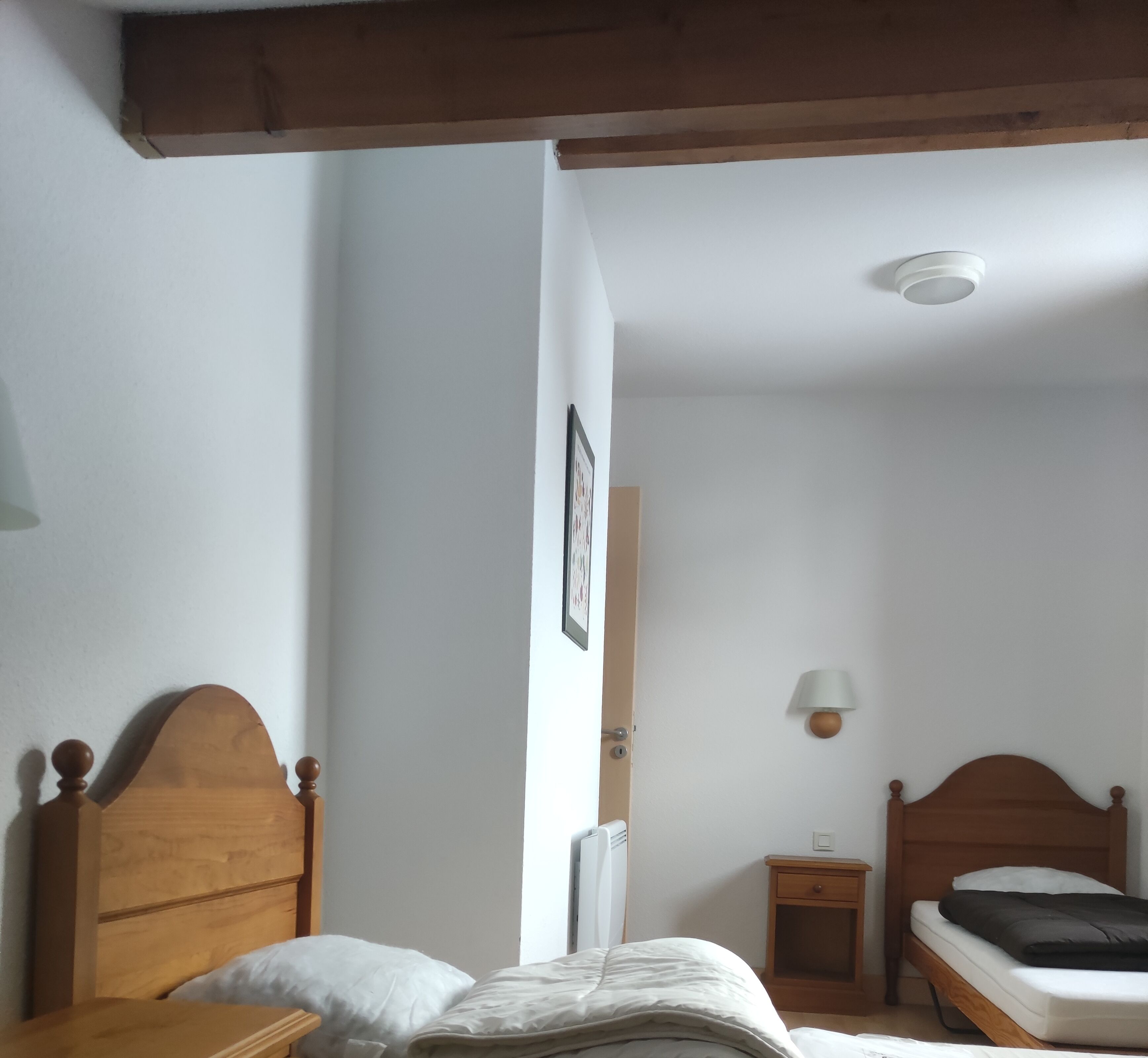 A bedroom with a wooden bed, a bedside table, a nightstand, a wall-mounted lamp, and a ceiling light.
