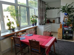 A compact kitchen with a dining area, featuring a window with potted plants, a red tablecloth, and wooden chairs.