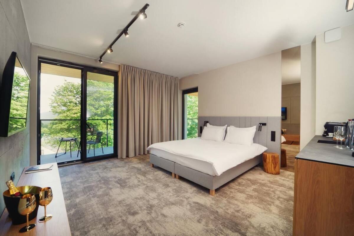 A modern bedroom with a large bed, a balcony with curtains, a desk with a chair, and a view of greenery outside.