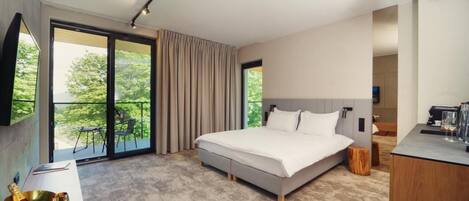 A modern bedroom with a large bed, a balcony with curtains, a desk with a chair, and a view of greenery outside.