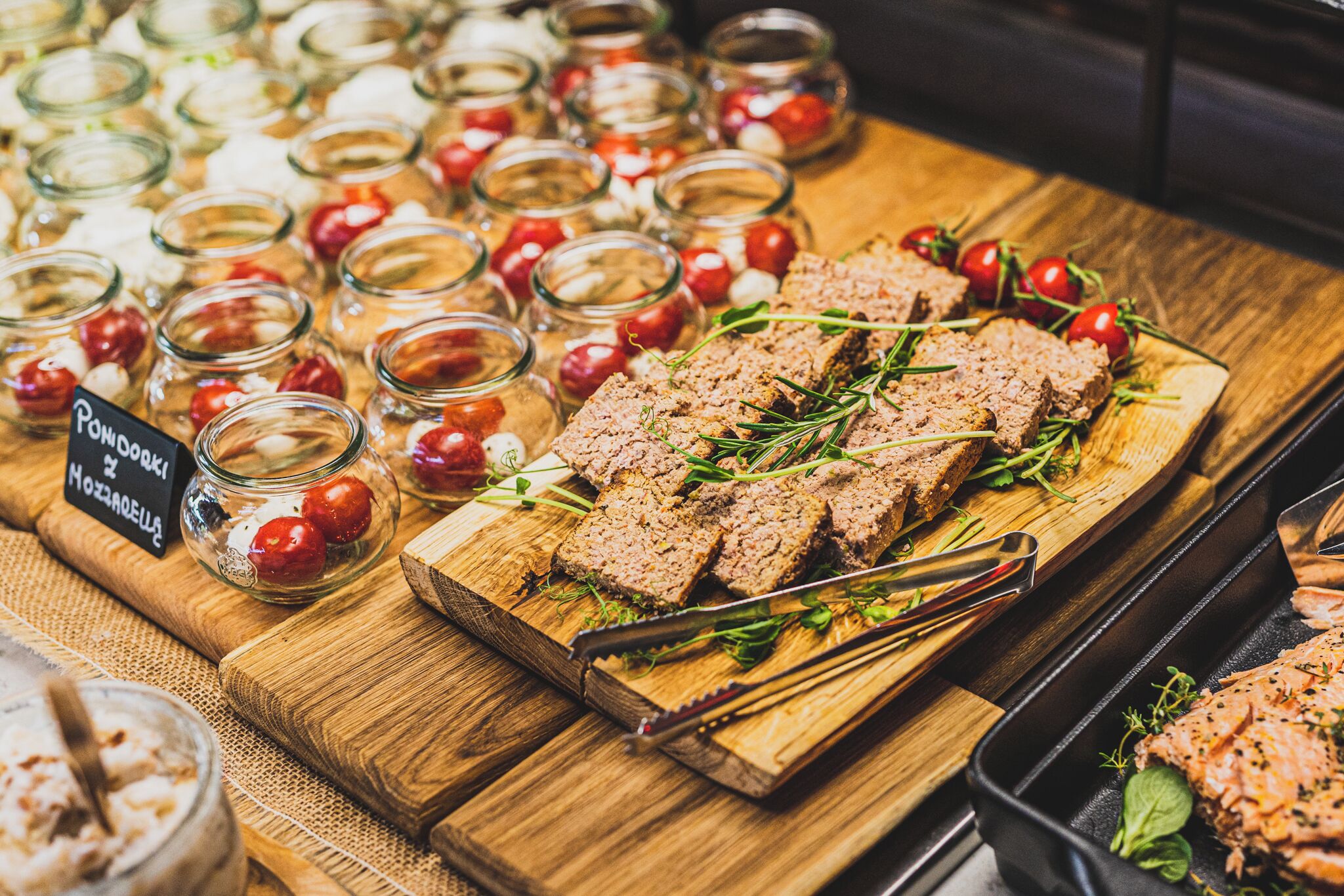 A wooden board with sliced meat, garnished with herbs, and surrounded by small glass jars with cherry tomatoes.