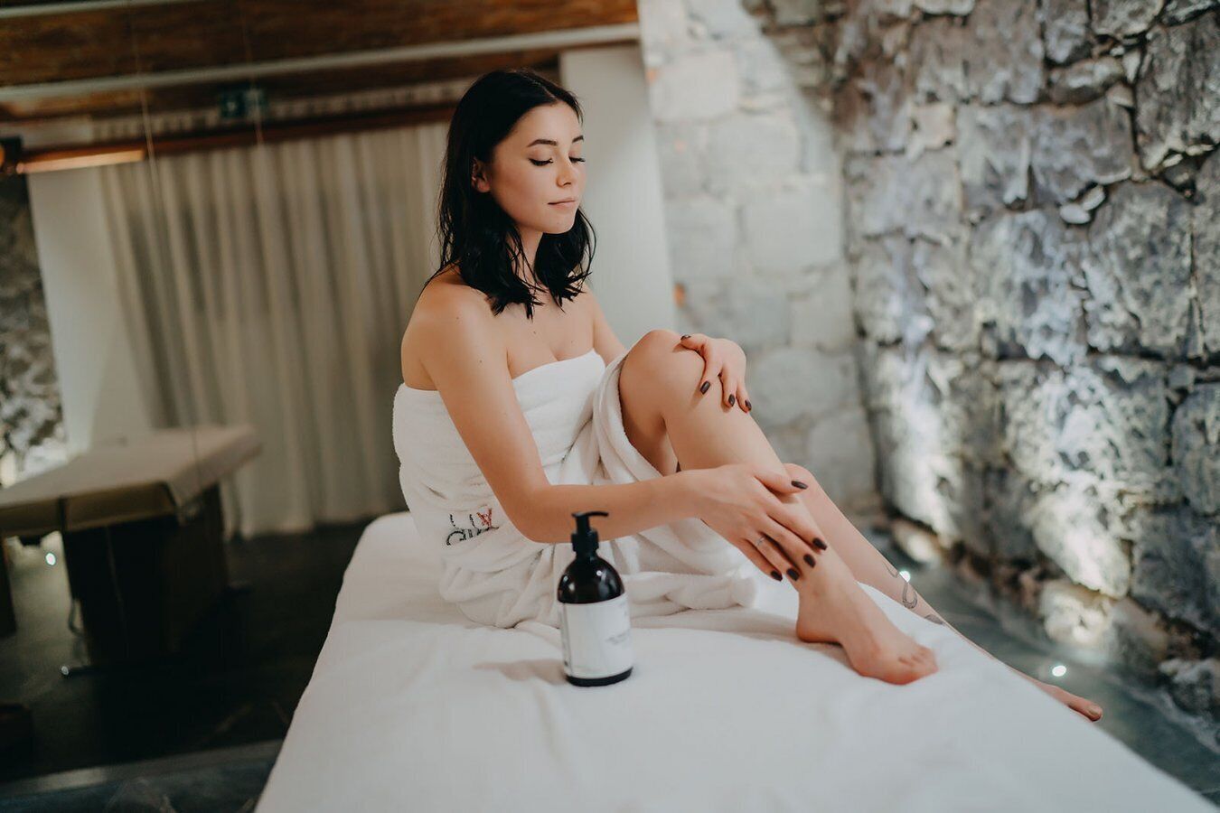 A person sitting on a bed in a spa-like setting with a stone wall and a massage table in the background.