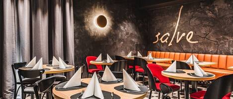 Un interior de restaurante con mesas redondas, sillas negras y rojas, y un piso a cuadros.