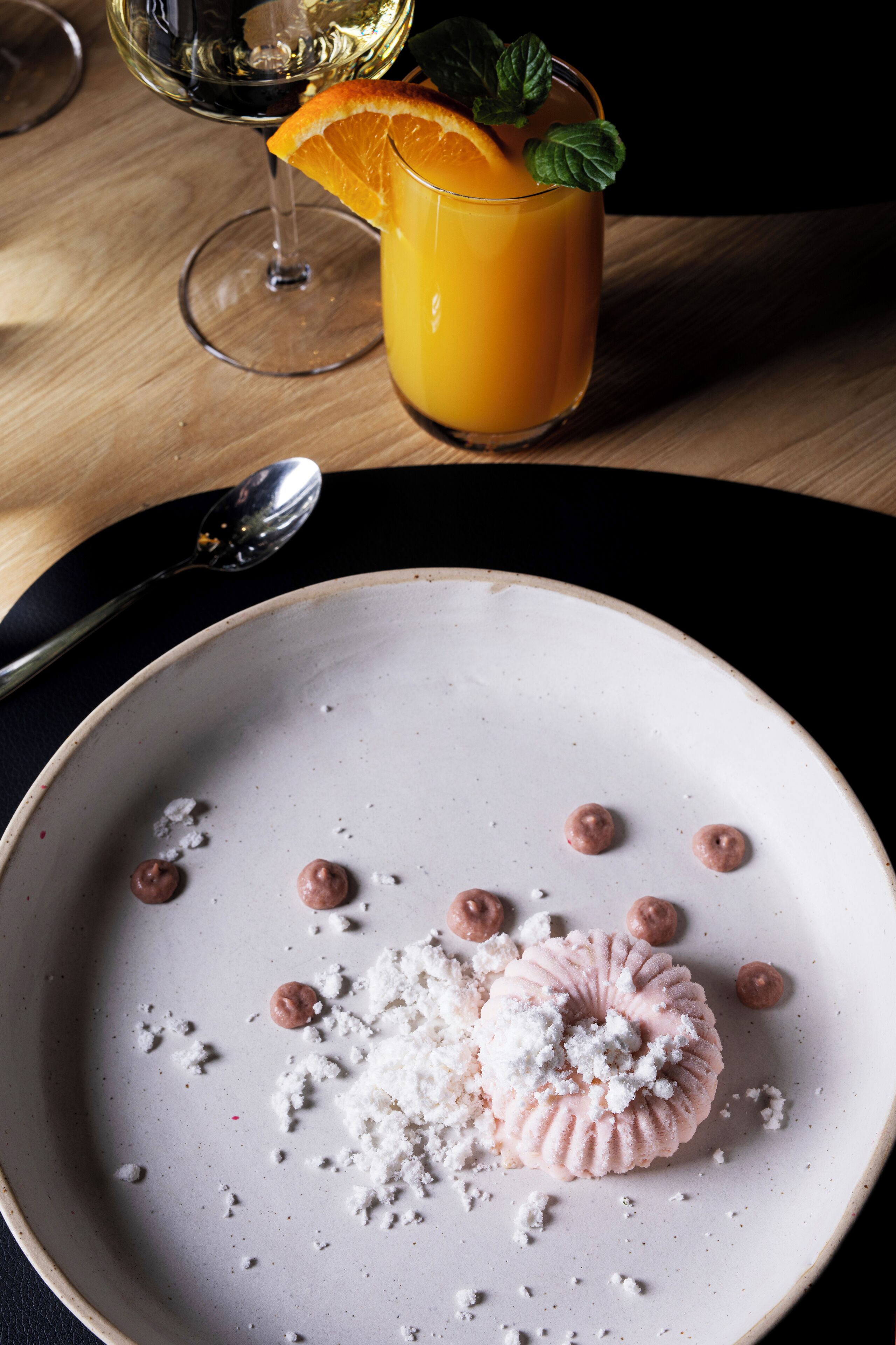 Un plato de postre con un dulce rosado, un vaso de jugo de naranja y una copa de vino sobre una mesa de madera.