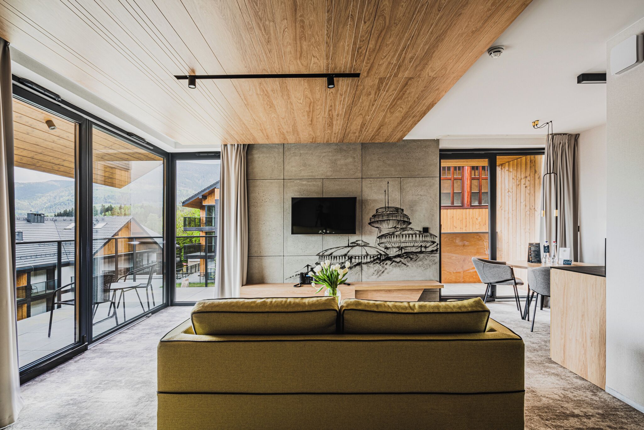 A modern living room with a large window, a flat-screen TV mounted on a concrete wall, and a dining area with a wooden table and chairs.