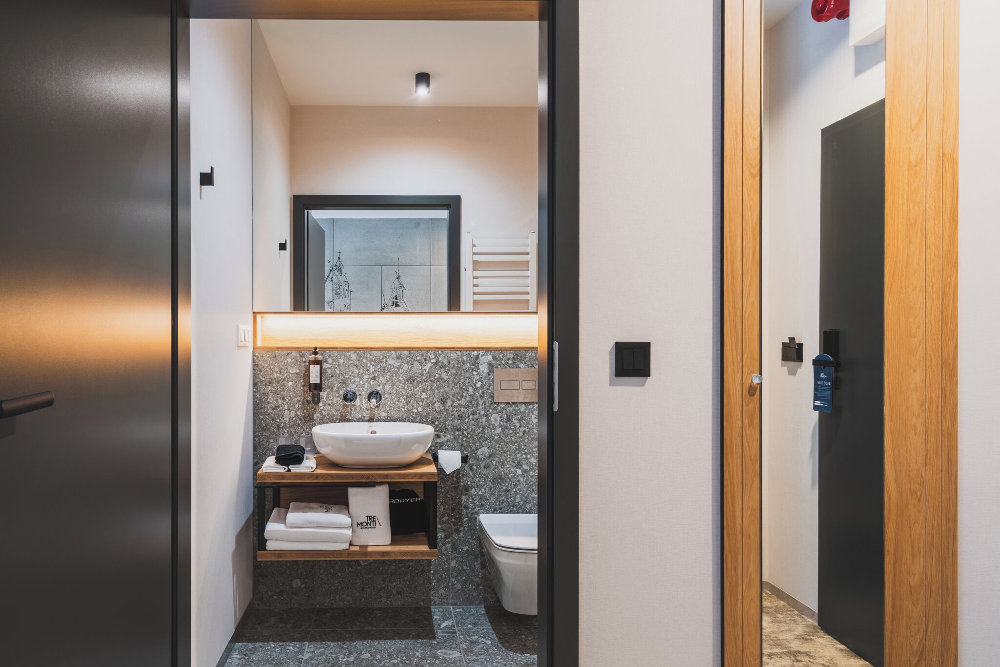 A modern bathroom with a large sink, a mirror, and a toilet. There is a towel rack and a shelf with towels.