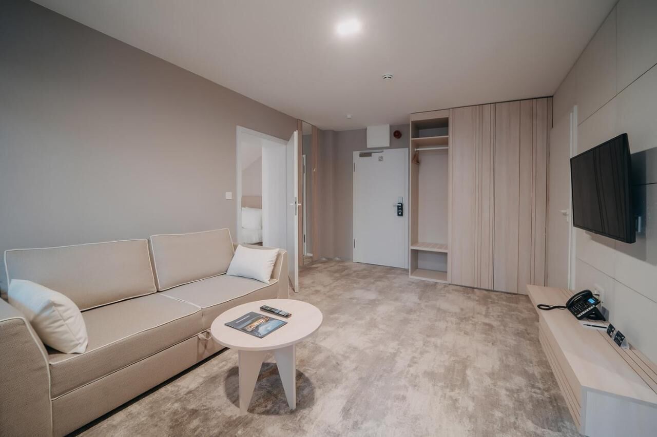 Una habitación de hotel moderna con un sofá color beige, una mesita redonda pequeña, un televisor montado en la pared y un vestidor.