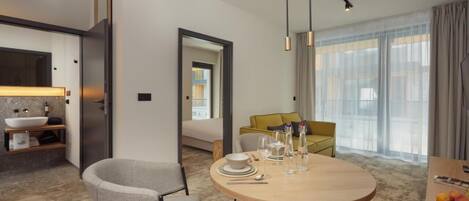Una habitación de hotel moderna con una mesa redonda de comedor, un sofá y un ventanal con cortinas.