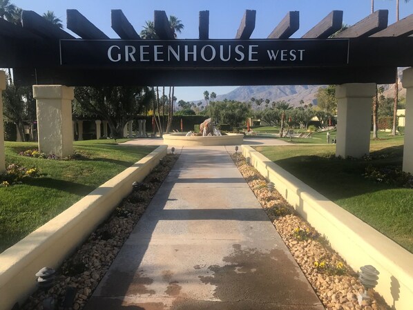 Uma trilha leva a um edifício com a placa "GREENHOUSE WEST", apresentando um lago central, palmeiras e montanhas ao fundo.