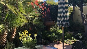 Une table ronde en verre, agrémentée d’une plante en pot, d’un parasol rayé et d’une chaise noire, dans un espace extérieur ensoleillé où l’on trouve diverses plantes et un mur en pierre.