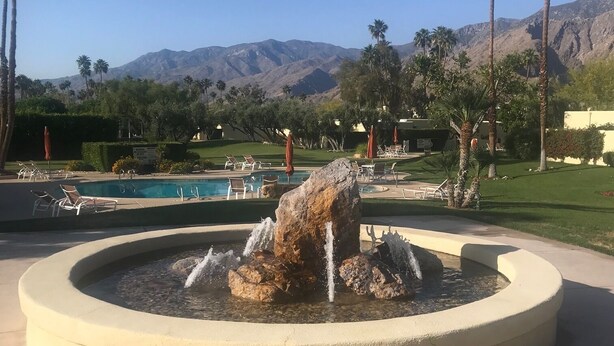 Une fontaine circulaire agrémentée de rochers et de jets d’eau, entourée d’une zone pavée avec des chaises longues et des parasols. Au fond, on aperçoit une piscine et un paysage montagneux parsemé de palmiers.