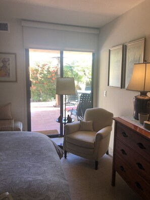 Ein Hotelzimmer mit Bett, Sessel, Kommode und Lampe. Ein Fenster bietet Blick auf einen Garten mit Gartenmöbeln.