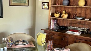 Un coin repas avec une table en verre dressée pour quatre personnes, entourée de chaises en osier. Un buffet en bois, orné d’objets décoratifs et de livres, se trouve à droite.