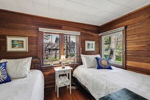 A room with two beds, wooden walls, a window with blinds, and a small table with a lamp.