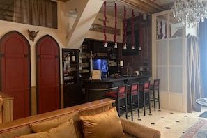 Een bar met een toonbank en krukken, een bank met een kussen, en een kroonluchter.