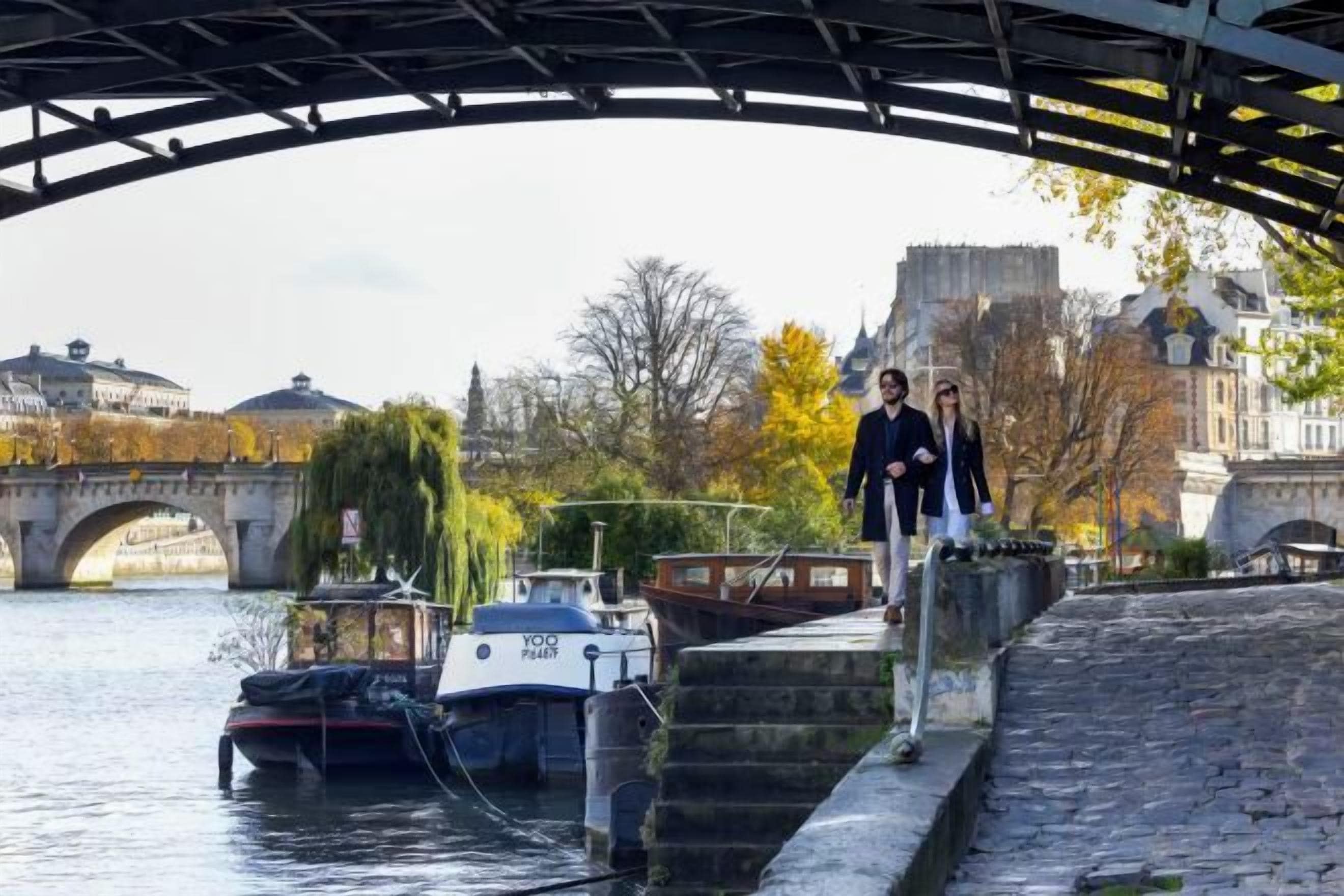 A bridge over a river with boats docked, a stone wall, and buildings in the background.