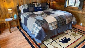 Quarto aconchegante com parede de madeira, cama com colcha xadrez, placa de boas-vindas e uma luminária.