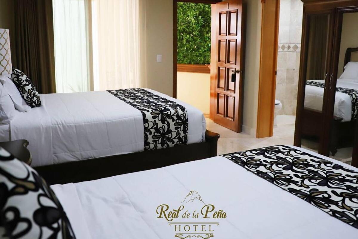 A hotel room with two beds, a large window with curtains, a wooden door, and a view of greenery outside.