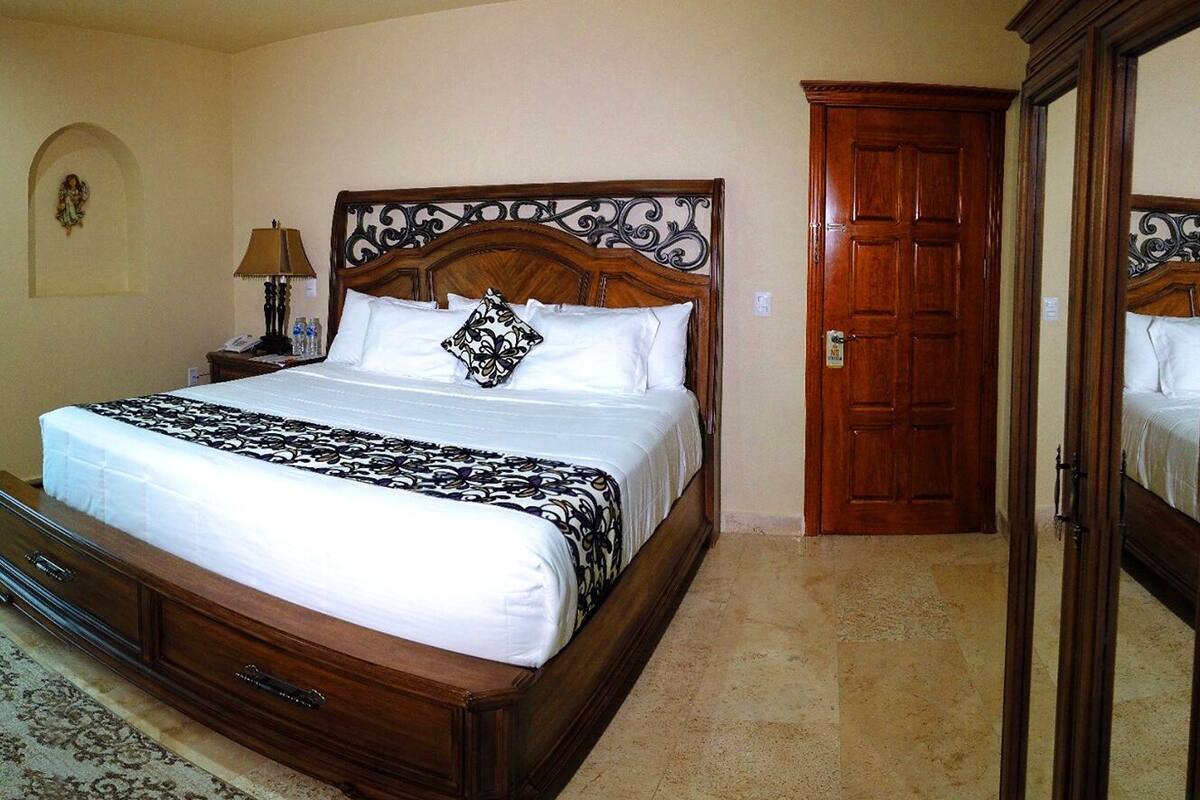 A hotel room with a large bed, a wooden wardrobe, a nightstand with a lamp, and a wall-mounted light fixture.