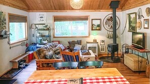 A cozy cabin interior with a wooden ceiling, a fireplace, a dining area with a checkered tablecloth, and a large window.
